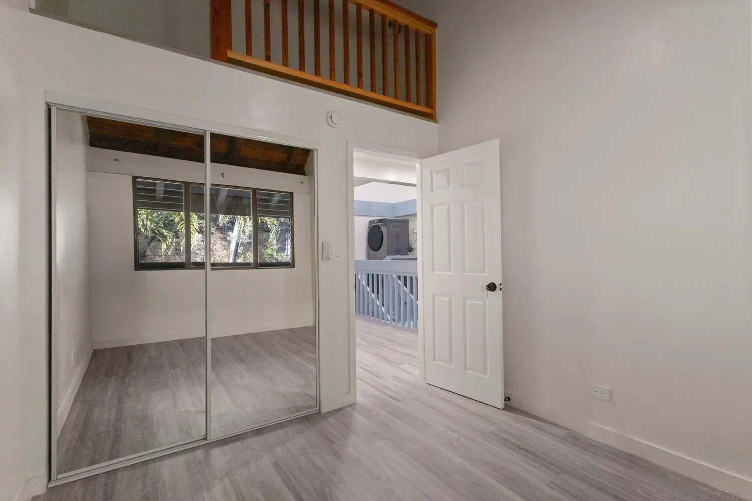 Bedroom with gray wood flooring, white walls, mirrored closet doors, and an open door leading to a balcony with laundry appliances outside.