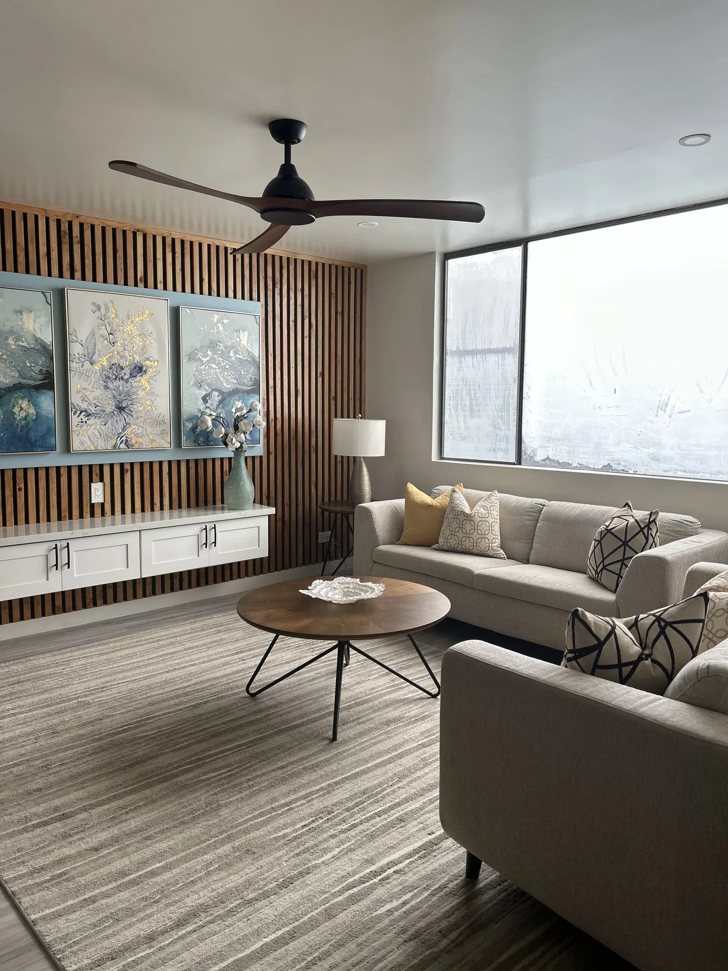 Living room with beige sofas, patterned cushions, round wooden coffee table, wooden wall panel, abstract artwork, side table with lamp, large window, ceiling fan, striped rug.