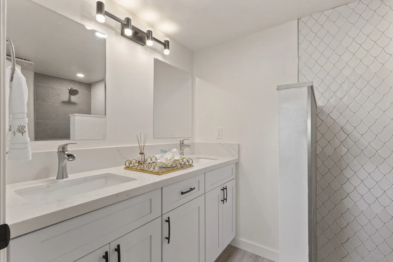 Modern bathroom with double vanity, white cabinets, marble countertops, and large mirror. There is a shower with gray tiles in the background and a decorative tile wall on the right. The lighting above the mirror has four cylindrical lights. A tray w