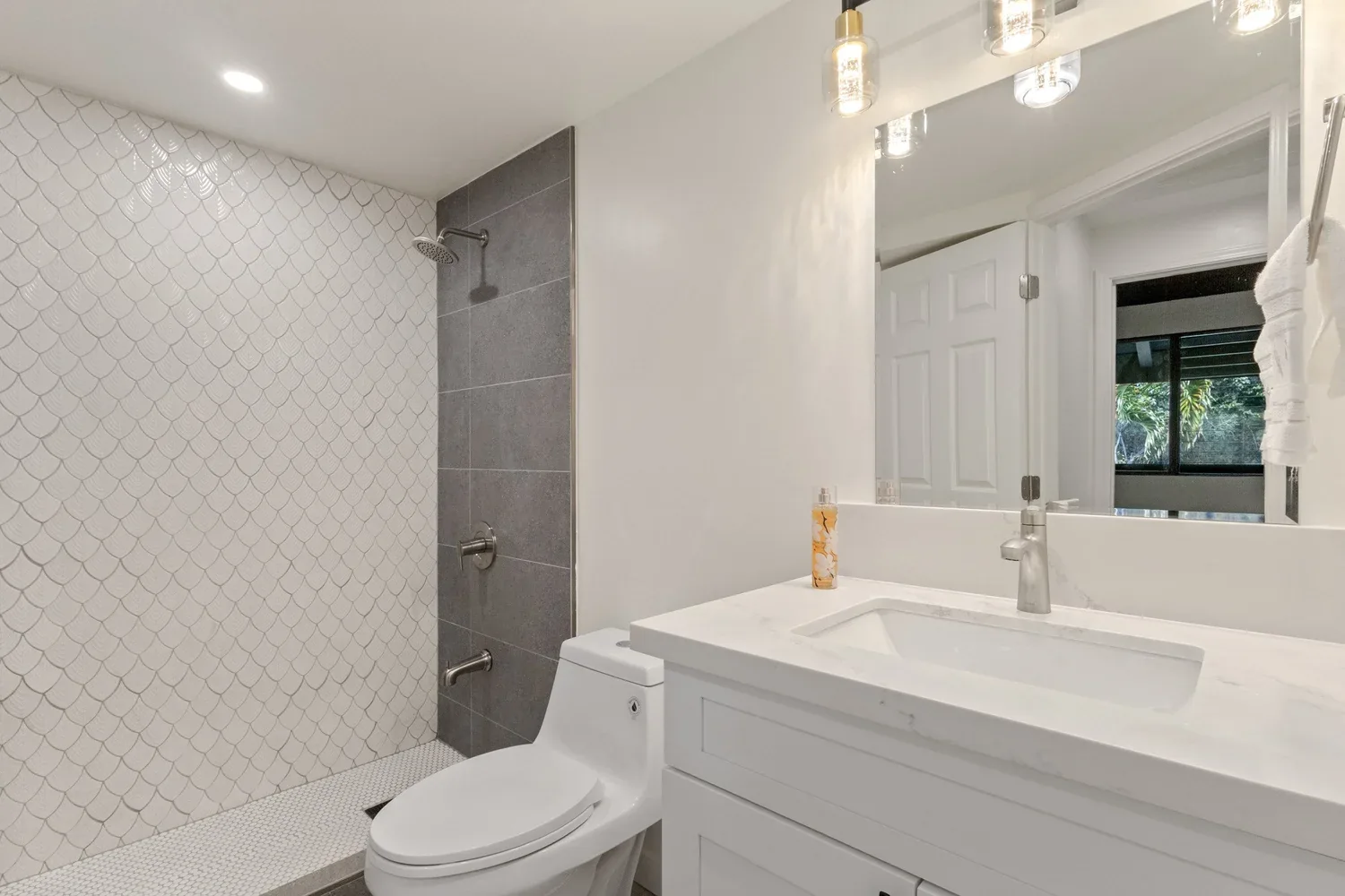 Modern bathroom with a walk-in shower featuring pebble tile floor and fish scale patterned wall, white vanity with marble countertop, large mirror, and a window showing greenery outside.