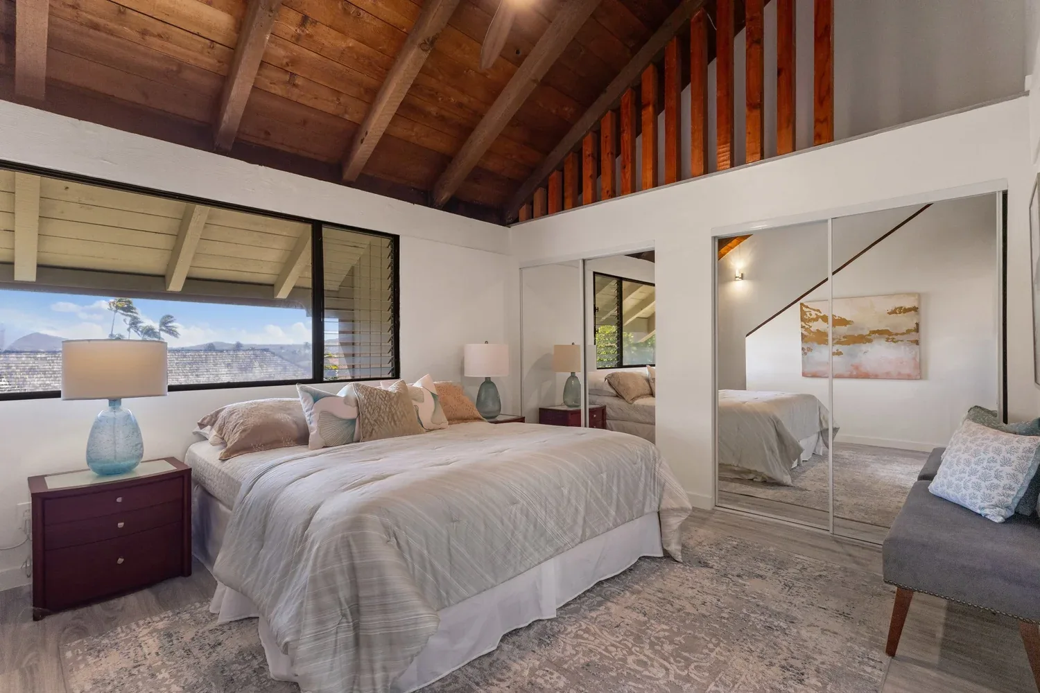 A bedroom with a large bed, nightstands with lamps, mirrored closet doors, and a grey sofa with pillows. The room has a high vaulted ceiling with exposed wooden beams, and large windows showing an outdoor view.