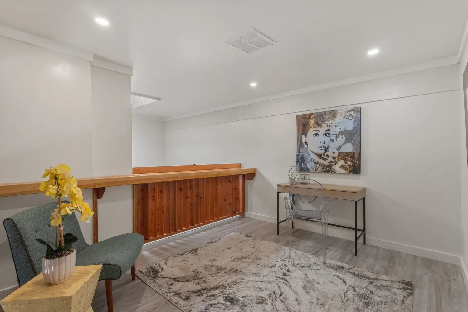 A room with white walls and ceiling, recessed lighting, a wooden railing, a desk with a transparent chair, a piece of artwork of a woman on the wall, a small table with a potted yellow orchid, and a patterned area rug on luxury vinyl flooring.