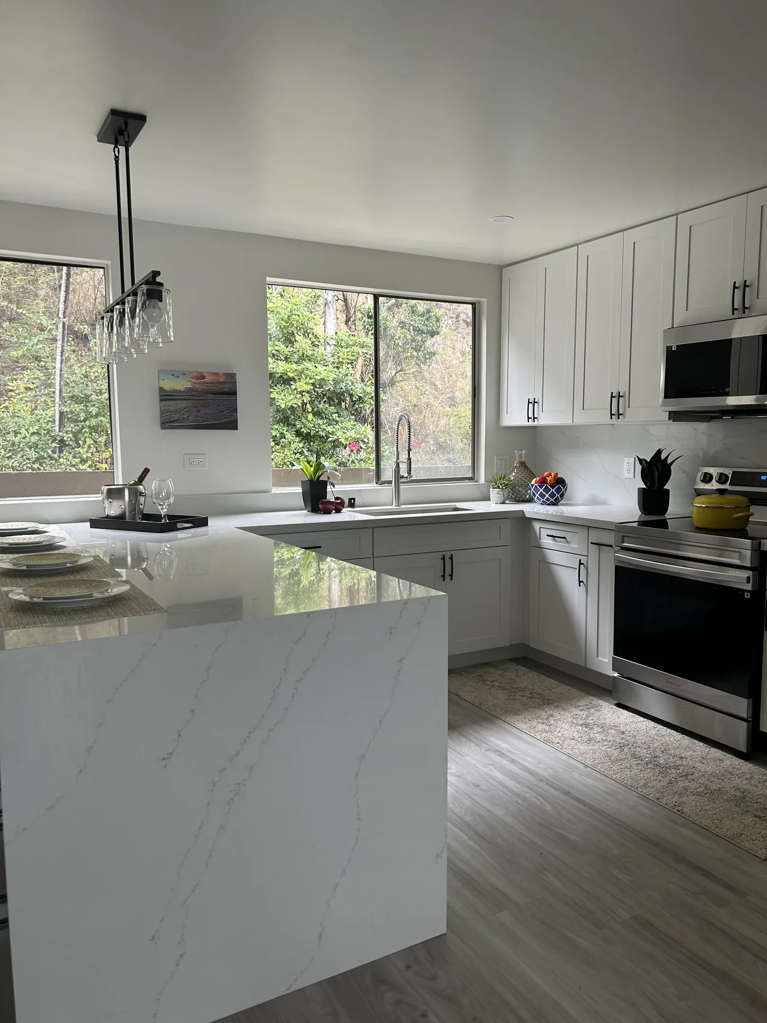Modern white kitchen with marble countertops, black hardware, and stainless steel appliances. Large window offers a view of greenery outside.